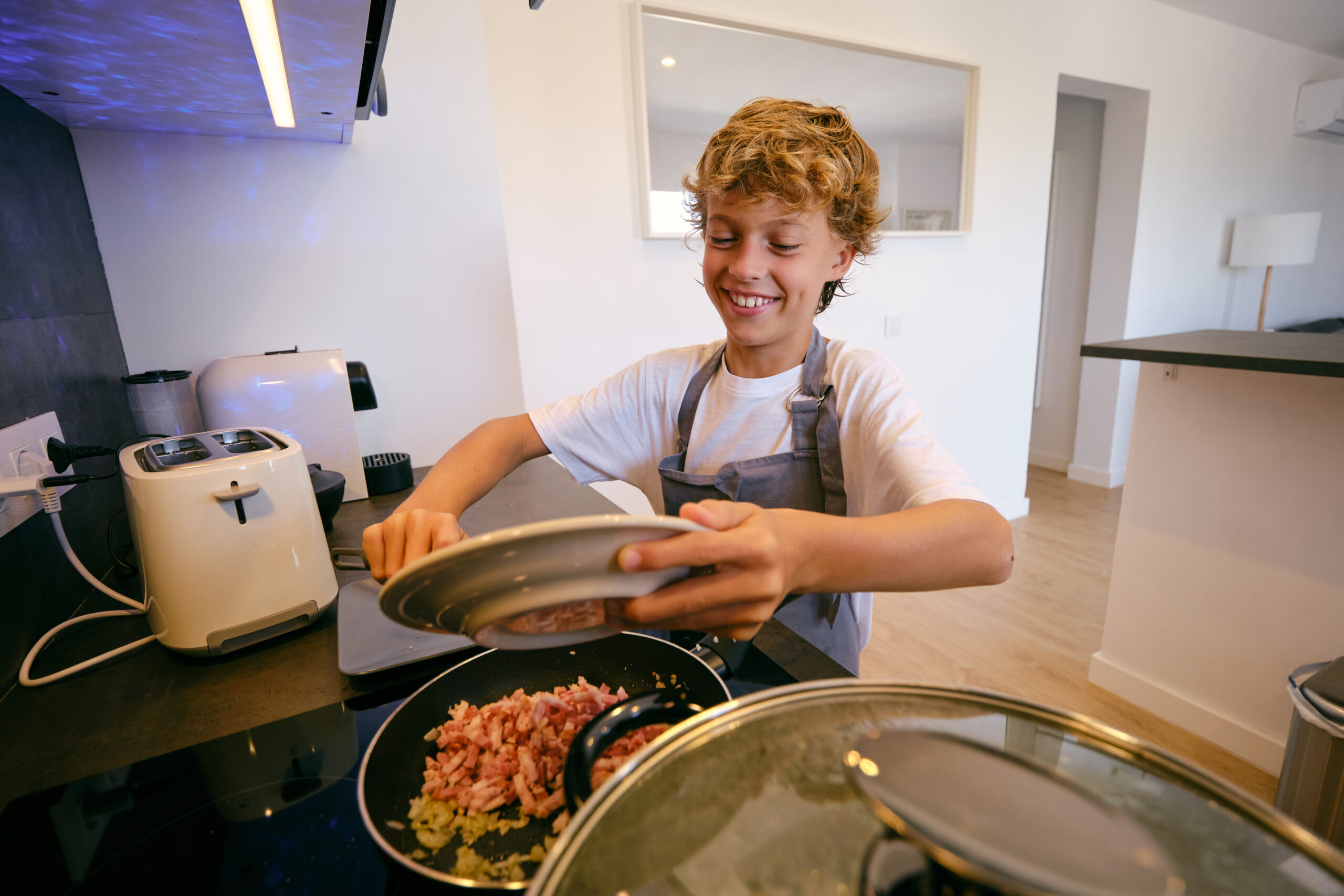 Cheerful child with plate putting smoked bacon in frying pan while cooking in house kitchen